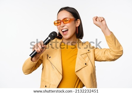 Stylish asian girl in sunglasses, singing songs with microphone, holding mic and dancing at karaoke, posing against white background Royalty-Free Stock Photo #2132232451