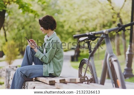SIde view happy young woman 20s in green jacket jeans sit on bench near bicycle in city spring park outdoors hold mobile cell phone chat online in social network People active urban youthful concept. Royalty-Free Stock Photo #2132326613
