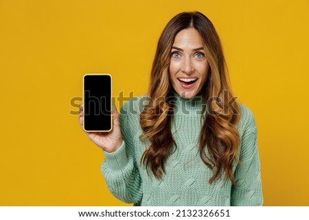Young smiling amazed woman 30s in green knitted sweater hold use mobile cell phone with blank screen workspace area isolated on plain yellow color background studio portrait. People lifestyle concept