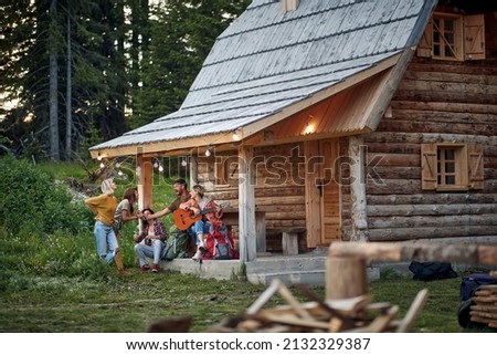 Young cheerful friends in front of wooden cottage on the terrace.  Girl playing guitar. Summertime garden celebration and fun. Friends, togetherness, fun, celebration concept. Royalty-Free Stock Photo #2132329387