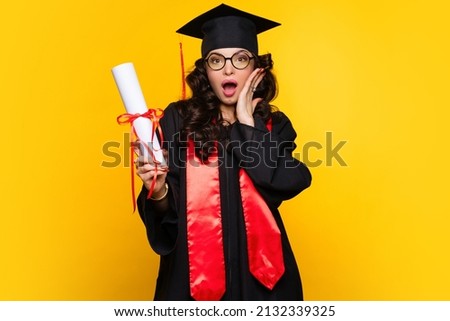 Shocked Girl graduate in graduation hat and eyewear with diploma on yellow backdrop. pleasantly surprised young woman wearing graduation cap and ceremony robe holding Certificate. Education Concept Royalty-Free Stock Photo #2132339325