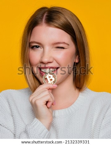 Girl biting btc coin and blinks eye. Woman holding golden bitcoin winking looking at camera. Advertise of digital money btc coin for e-commerce and nft projects. Try cryptocurrency and blockchain. Royalty-Free Stock Photo #2132339331