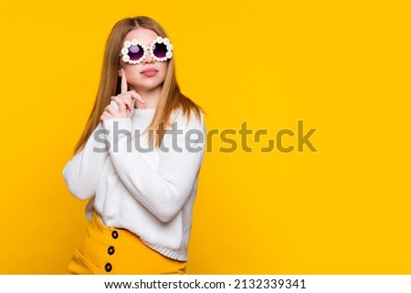Attractive girl wears white sweater and stylish daisies sunglasses keeps hands together looks away with dreamy expression dreams. Young woman wait oncoming of spring on yellow background. Lifestyle Royalty-Free Stock Photo #2132339341