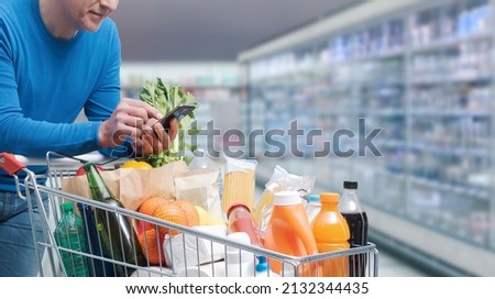 Man doing grocery shopping at the supermarket, he is pushing a trolley and using a smartphone