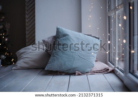 pillows in cold shades lie on a wide white window sill, the window is decorated with garlands