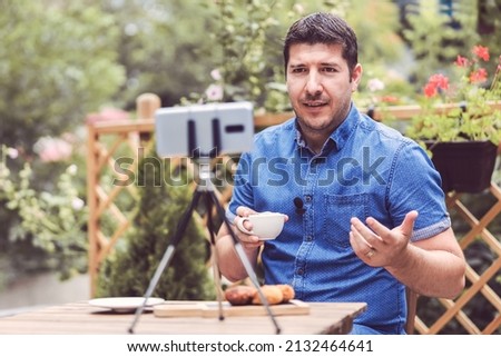 Social media influencer or food blogger creating content at restaurant terrace table - man with lavalier sharing online food review using smartphone on tripod - content creator vlogger filming video