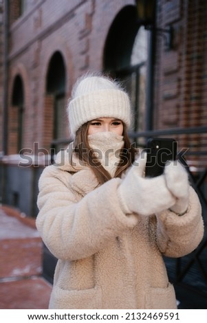 A young woman dressed warmly in winter clothes takes a photo with her phone