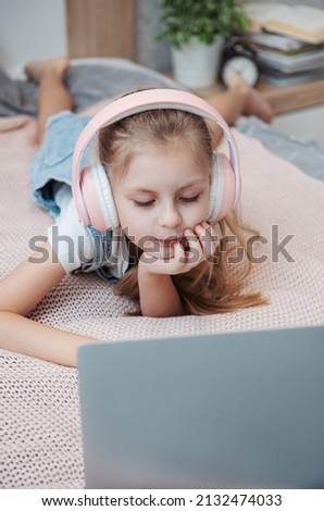 Smiling happy little kid girl using laptop in wireless headphones in bed at home.