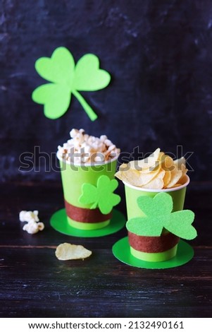 Popcorn and chips in green paper glasses in the shape of St. Patrick's hat, background decorated with shamrock leaves, on a wooden table, beer snack, concept congratulation, parties, postcard