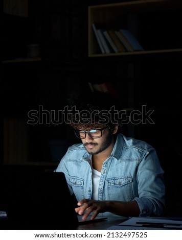 Young indian ethnic businessman developer or web designer wearing glasses typing on laptop distance working online late at night at home or in dark office using laptop sitting at workplace desk. Royalty-Free Stock Photo #2132497525