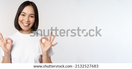 Very well, excellent. Smiling asian woman showing okay sign, approval, ok gesture, looking satisfied, recommending smth, standing over white background