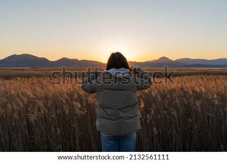 A woman standing in a reed field holds off her ears as she looks at the sunset