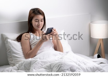 Beautiful young Asian woman relax in bedroom wake up in the morning texting messaging on phone.