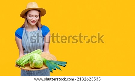 Woman farmer gardener wearing straw hat apron holding vegetables cabbage celery and leek on wooden tray on yellow background