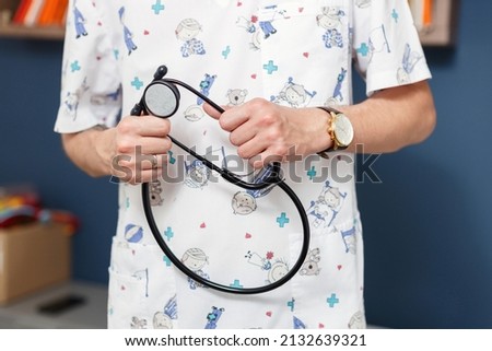 married young male pediatrician in a white coat with cartoon childish characters holding a fenendoscope in his hands, close-up without a face