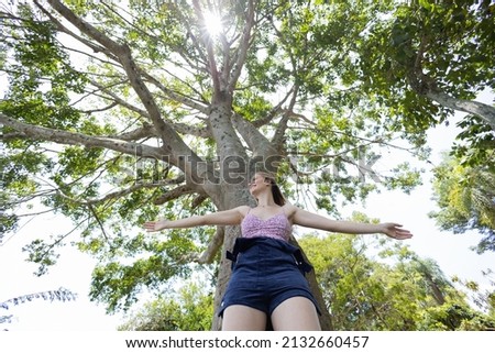 Portrait of smiling young woman at a park on a beautiful sunny day.  Woman with open arms in front of a big a tree. Green and nature background. High quality photo