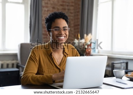 Joyful happy short haired Black business woman in glasses sitting at workplace with laptop in home office, looking at camera, smiling. Millennial worker, employee, entrepreneur head shot portrait