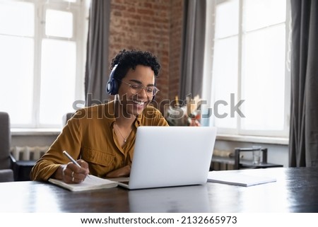 Cheerful Black college student in earphones attending virtual class, writing notes, watching webinar on Internet, studying online. African worker in headphones talking to customer on video call Royalty-Free Stock Photo #2132665973