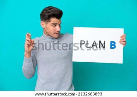 Young handsome caucasian man isolated on blue bakcground holding a placard with the message PLAN B with fingers crossing