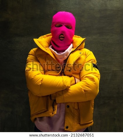 Half-length portrait of young anonymous man wearing pink balaclava and yellow down jacket, coat isolated on dark vintage background. Concept of safety, art, fashion. Royalty-Free Stock Photo #2132687051