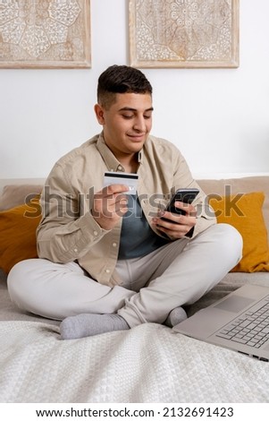 Young man smiling while surfing the web on his smartphone and holding credit card in his hand trying to buy something online. Easy online shopping at home, e-commerce. Cosy interior.