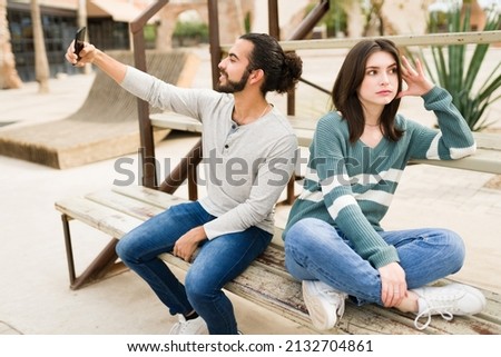 Bored attractive woman looking tired while her boyfriend uses a smartphone to record a short story for social media