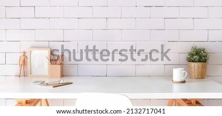 Stylish workplace with picture frame, coffee cup, potted plant and supplies on white table. Copy space.