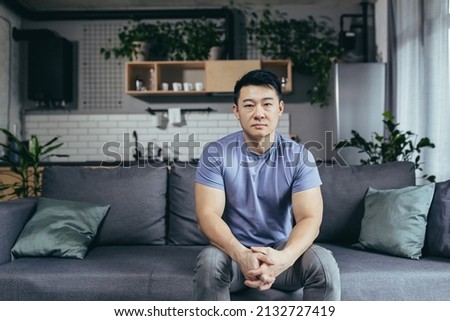 Portrait of a serious man at home, Asian man sitting on the couch looking intently at the camera