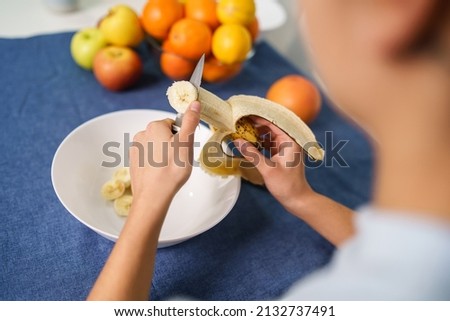 Close up on hands of unknown caucasian woman cutting banana by the table at home - back view top angle copy space - healthy eating concept