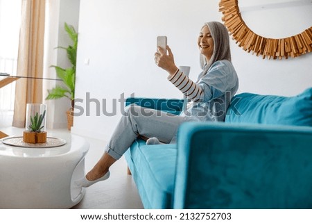 Cheerful female person holding cup of hot drink and taking picture with smartphone while sitting on couch