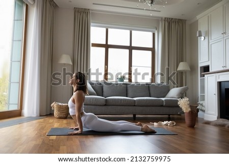 Young sporty yogini woman in fashion comfy white sportswear practicing yoga asanas at home, doing Upward Facing Dog exercise, full-length. Strengthens back muscles, lengthens spine. Training concept Royalty-Free Stock Photo #2132759975