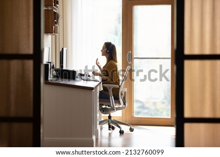 In modern homeoffice young business woman sit on office chair wear headphones talks on video conferencing, foreign language teacher lead on-line class, side view. Videocall application usage concept Royalty-Free Stock Photo #2132760099