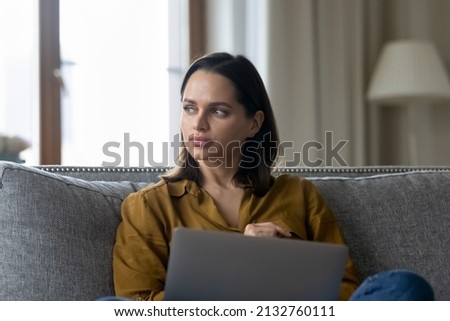 Young serious freelancer woman sit on couch with laptop, ponder, search solution, thinks over issue, decide business in quarantine at home looks aside. Blogging, new ideas searching, telework concept