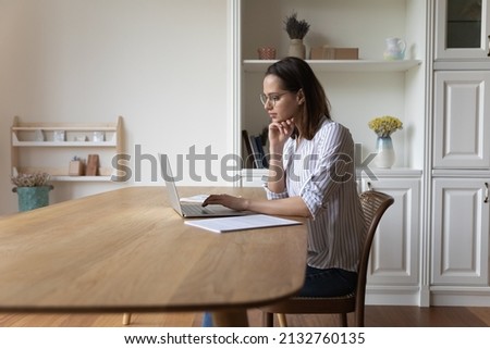 Young woman in glasses sit at table looks at laptop screen makes research use internet, web surfing information, do assignment studying or working online use wireless computer. Tech, freelance concept