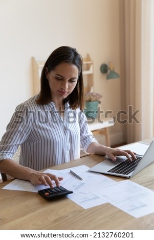Vertical image serious housewife sit at table at home kitchen check household bills, make calculations on calculator, pay utilities through e-banking system on laptop. Manage family finances concept