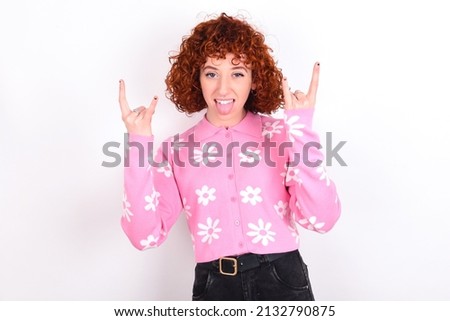 young redhead girl wearing pink floral t-shirt over white background making rock hand gesture and showing tongue