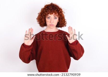 Serious young redhead girl wearing red sweater over white background pulls palms towards camera, makes stop gesture, asks to control your emotions and not be nervous
