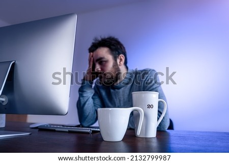 Tired man sits in front of a computer with a cup of coffee, colored lighting.
