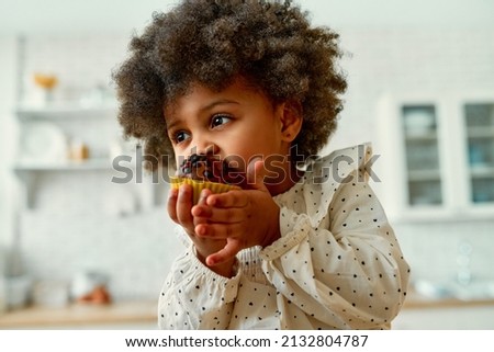 An African American little baby with curly fluffy hair in a dress eats a chocolate muffin with appetite in the kitchen at home. Royalty-Free Stock Photo #2132804787