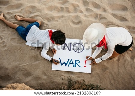 top view of a Caucasian mother and her Asian son writing on a sign the phrase no war. they are on the beach at sunset. no war and peace in the world concept.