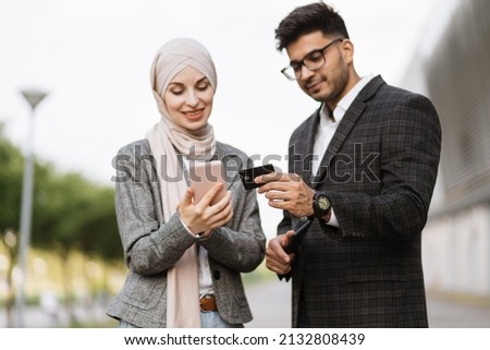 Conceptual photo of hindu businessman showing credit card number to his female Muslim colleague typing on smartphone and making online payment. Happy people outdoors shopping online.
