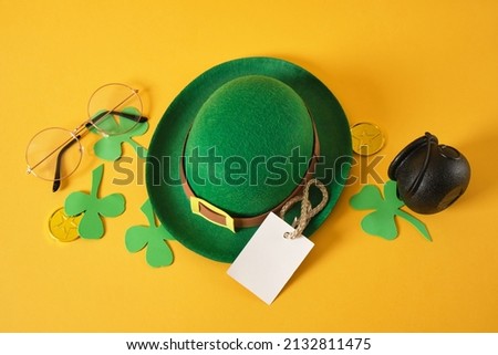white blank tag, glasses, cauldron, gold coins, decorative green leprechaun felt hat from and leaves from foamiran on a yellow background, st. patrick's day concept, copy space