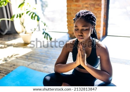 african american woman in black sporty bra is sitting lotus pose on yoga mat floor closed eyes and listening an audio meditate mantra hands namaste Royalty-Free Stock Photo #2132813847