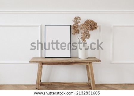 Vase with dry hydrangea flowers on old wooden bench. Blank black picture frame mockup. White wall moulding background, stucco decor. Empty copy space. Elegant interior. Summer, fall still life photo. 