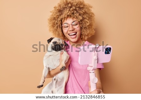Cute cheerful female pet owner poses with pug dog for selfie spend free time together make memorable photo while having walk isolated over brown background. Glad young woman with domestic animal
