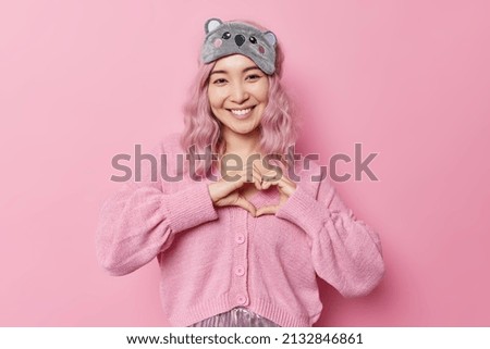 Positive young pink haired Asian girl makes heart gesture expresses love smiles pleasantly wears sleepmask on forehead and casual jumper isolated over pink background. Body language concept.
