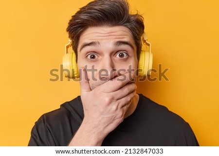 Shocked young Caucasian man covers mouth with hand tries to be speechless stares bugged eyes listens music via stereo headphones cannot believe in shocking news isolated over yellow background