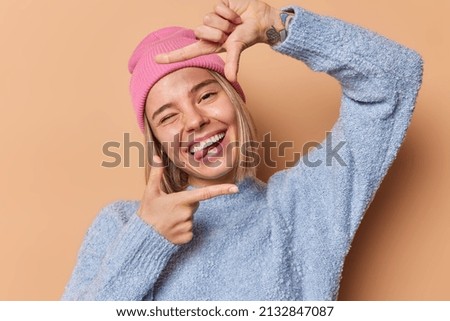 Beautiful young woman makes frame gesture over head winks eye sticks out tongue searches perfect angle makes picture has playful mood wears casual jumper and hat isolated over brown background