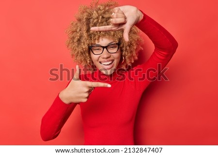 Positive curly haired woman searches perfect angle takes photo makes frame gesture as if photographing with camera smiles positively wears spectacles and turtleneck isolated on red studio wall