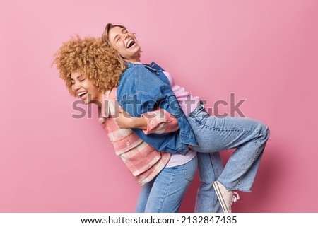 Two funny beautiful women give piggy back to each other have fun and laugh happily dressed in fashionable clothes isolated over pink background spend free time together havent seen for long time Royalty-Free Stock Photo #2132847435